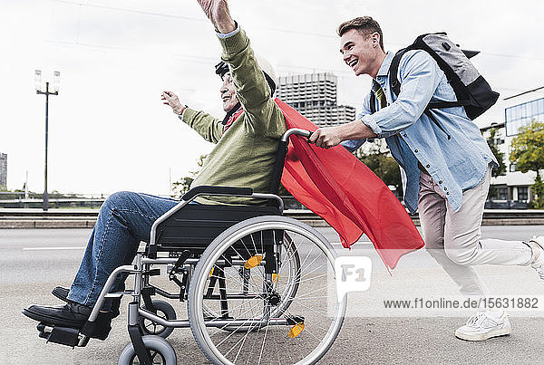 Junger Mann schiebt älteren Mann  der als Superheld verkleidet in einem Rollstuhl sitzt