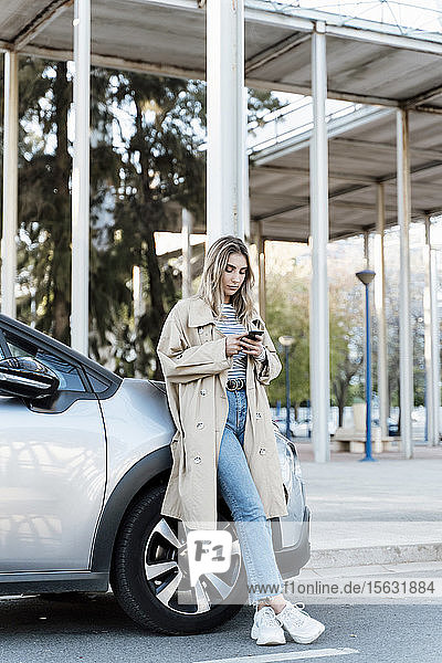 Young blond woman using smartphone  leaning on a car
