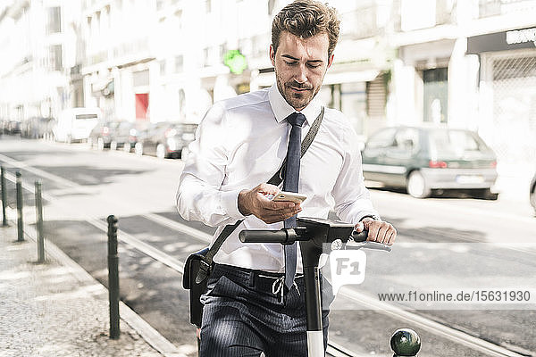 Junger Geschäftsmann mit einem E-Scooter  der ein Mobiltelefon in der Stadt benutzt  Lissabon  Portugal