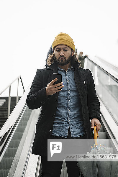 Porträt eines Mannes mit Regenschirm  der auf der Rolltreppe steht und auf ein Smartphone schaut