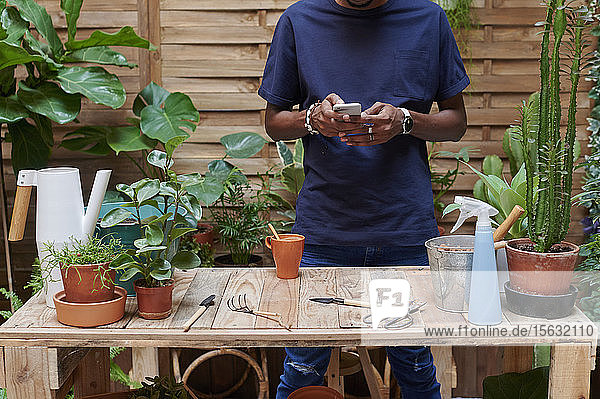 Junger Mann benutzt Smartphone auf seiner Terrasse bei der Gartenarbeit