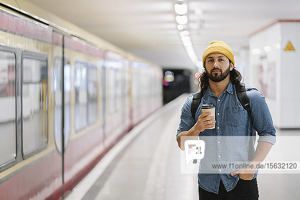 Porträt eines Mannes mit Rucksack und Kaffee zum Mitnehmen beim Warten am Bahnsteig  Berlin  Deutschland