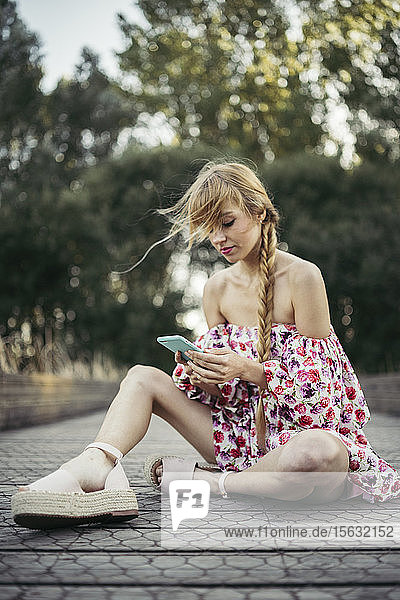 Porträt einer jungen Frau in Sommerkleid mit Blumenmuster  die mit ihrem Handy auf der Strandpromenade sitzt