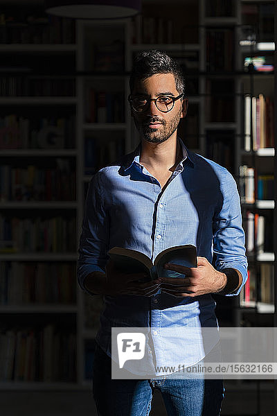 Porträt eines jungen Mannes mit Buch  der zu Hause vor einem Bücherregal steht