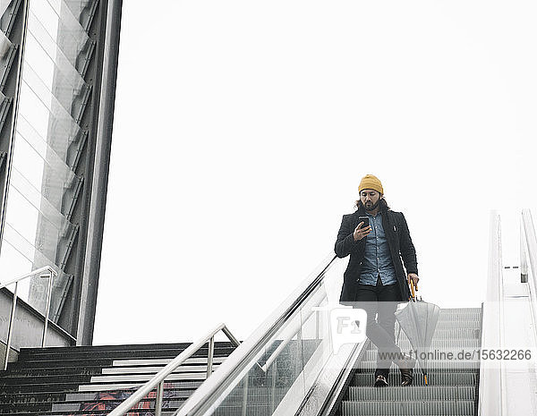 Mann mit Regenschirm steht auf Rolltreppe und schaut auf Smartphone  Berlin  Deutschland