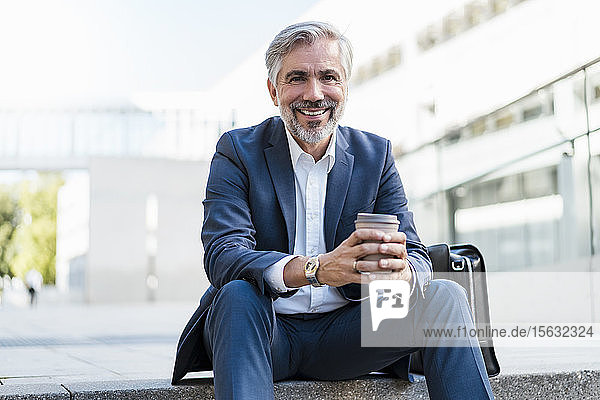 Porträt eines lächelnden reifen Geschäftsmannes  der auf einer Treppe in der Stadt sitzt und Kaffee zum Mitnehmen in der Hand hält