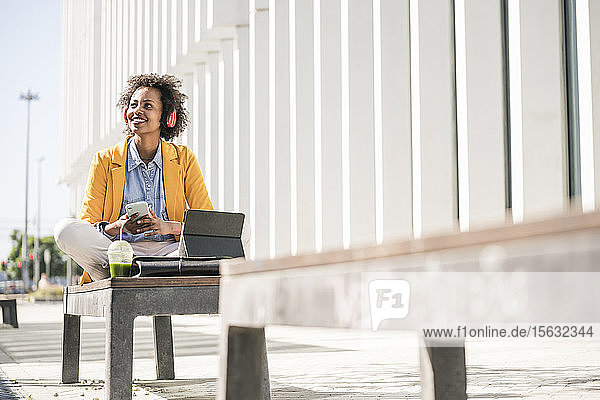 Smiling young woman with headphones  smartphone and tablet in the city