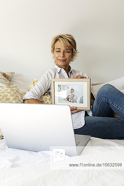 Reife Frau sitzt zu Hause auf dem Bett und zeigt ein Bild ihres Enkels auf einem Videoanruf