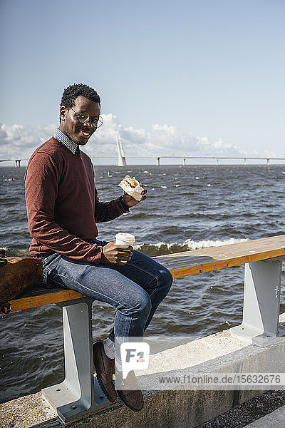Junger Mann sitzt an der Reling am Meer  isst Hamburger  trinkt Kaffee