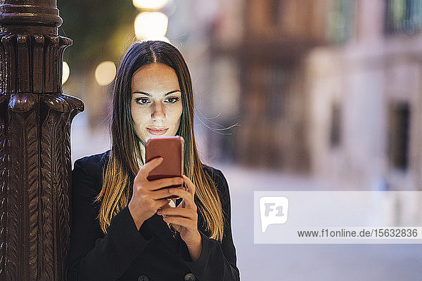 Porträt einer jungen Frau  die sich abends an einen Lampenmast lehnt und auf ein Handy schaut