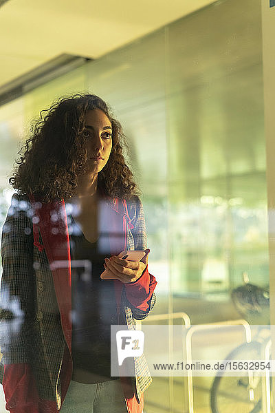 Porträt einer nachdenklichen jungen Frau mit Handy  die hinter einer Glasscheibe steht und in die Ferne schaut