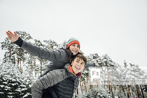 Porträt eines lächelnden jungen Mannes  der seine glückliche Freundin im Winterwald Huckepack nimmt