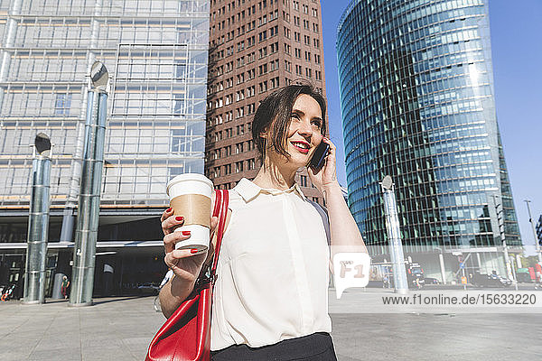 Lächelnde junge Geschäftsfrau mit Kaffee zum Mitnehmen am Telefon in der Stadt  Berlin  Deutschland
