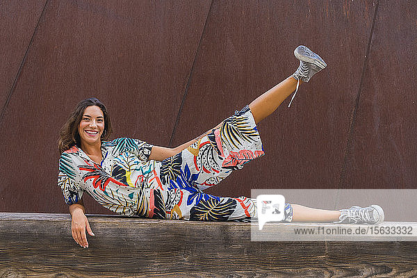 Porträt einer lächelnden jungen Frau  die auf einer Holzbank liegt und ihr Bein hebt