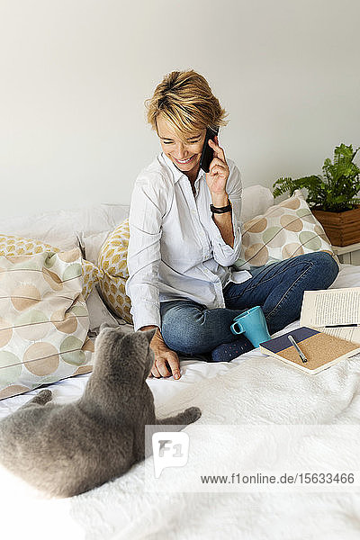 Reife Frau mit Katze sitzt zu Hause auf dem Bett und telefoniert