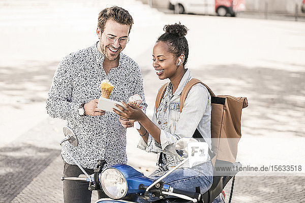 Glückliches junges Paar mit Motorroller und Eiscreme mit dem Handy in der Stadt  Lissabon  Portugal