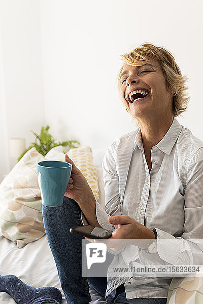 Lachende reife Frau sitzt zu Hause auf dem Bett und hält Smartphone und Kaffeetasse