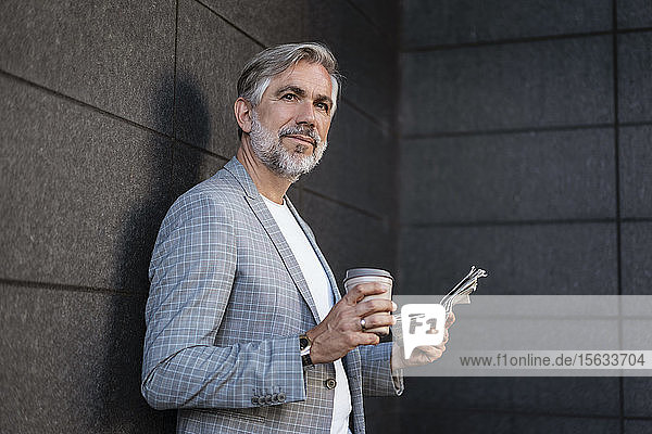 Modischer  reifer Geschäftsmann lehnt mit Zeitung und Kaffee zum Mitnehmen an einer Wand