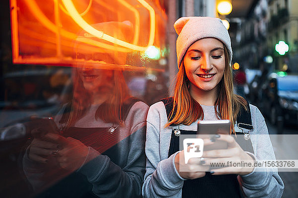 Lächelnde junge Frau steht nachts neben Neonlicht und benutzt ihr Smartphone