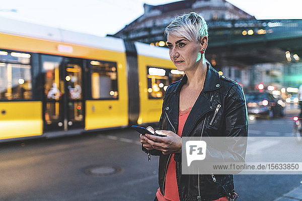 Porträt einer blonden Frau  die abends mit dem Handy am Straßenrand steht  Berlin  Deutschland