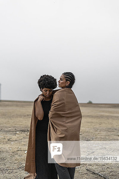 Two women standing in bleak landscape sharing blanket
