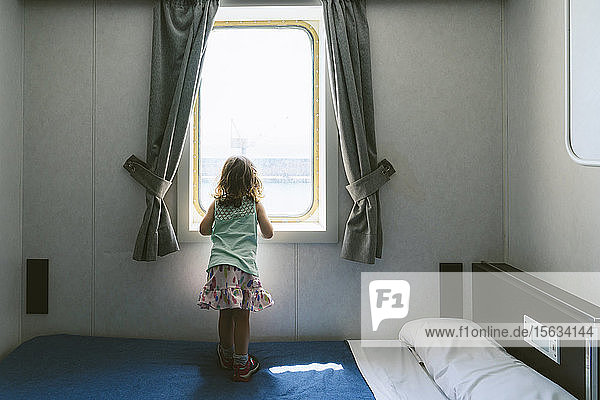 Rückenansicht eines kleinen Mädchens  das auf einem Bett steht und aus dem Fenster der Schiffskabine auf das Meer schaut