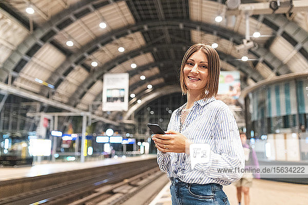Junge lächelnde Frau benutzt Smartphone am Bahnhof