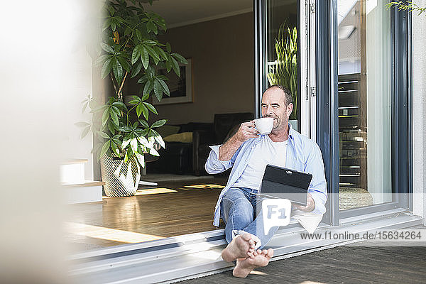 Mature man sitting at terrace door at home with tablet and coffee cup