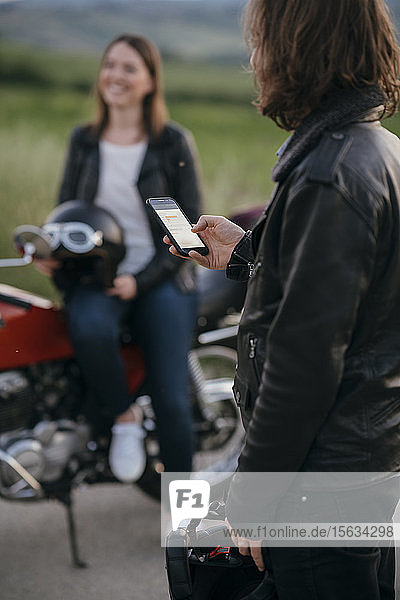 Junger Motorradfahrer steht mit seinem Smartphone auf der Landstraße  während seine Freundin im Hintergrund wartet