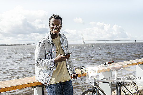 Junger Mann lehnt an der Reling am Meer und benutzt ein Smartphone