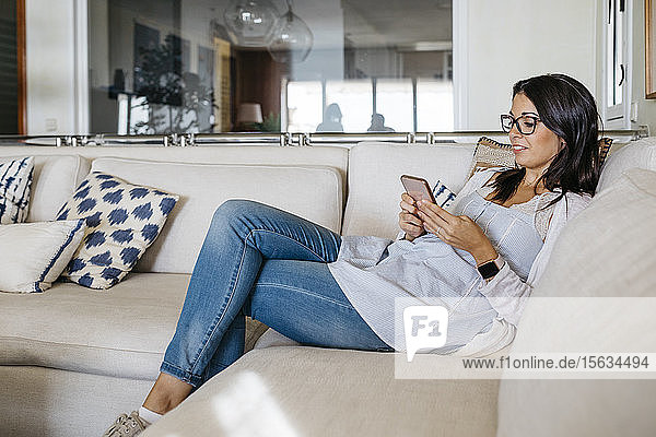 Lächelnde Frau zu Hause  die auf der Couch sitzt und auf ihr Handy schaut