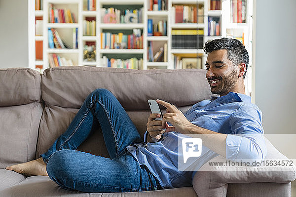 Lächelnder junger Mann liegt zu Hause auf der Couch und benutzt ein Smartphone