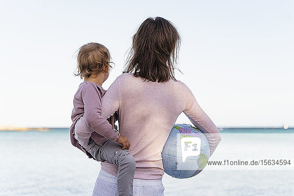 Rückenansicht einer Frau  die eine kleine Tochter hält  und eines Strandballs der Erde  der auf den Horizont schaut