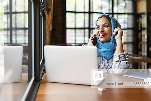 Geschäftsfrau  die in einem Café einen türkisfarbenen Hijab trägt und mit ihrem Laptop arbeitet  am Telefon