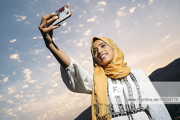 Junge muslimische Frau die einen gelben Hidschab trägt ein Smartphone benutzt und abends ein Selfie macht