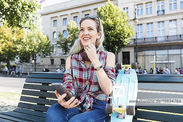 Porträt einer lächelnden jungen Frau auf einer Bank sitzend mit Skateboard und Mobiltelefon