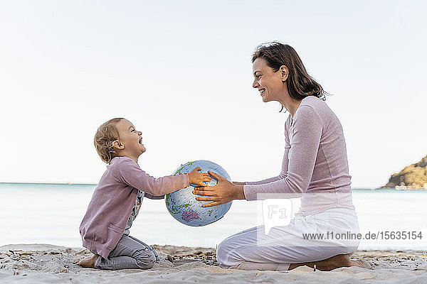 Glückliches kleines Mädchen sitzt mit seiner Mutter am Strand und spielt mit dem Earth Beachball