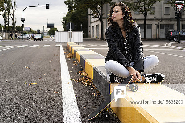 Junge Frau mit Skateboard sitzt auf einem Poller und schaut in die Ferne