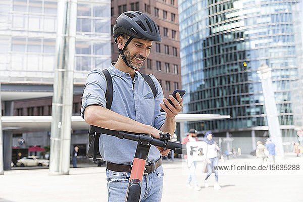 Smiling businessman looking at the smartphone commuting on electric scooter in the city  Berlin  Germany