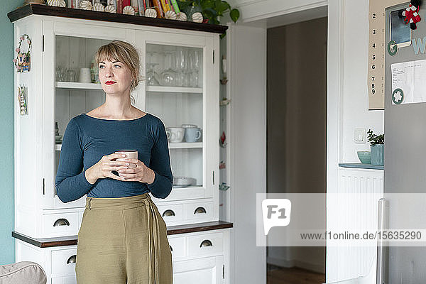 Porträt einer Frau  die mit einer Tasse Kaffee in der Küche steht und in die Ferne schaut
