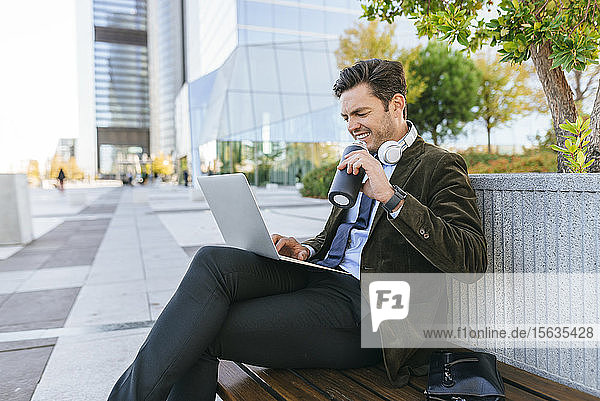 Smiling businessman sitting on bench in the city using laptop and drinking from refillable bottle  Madrid  Spain