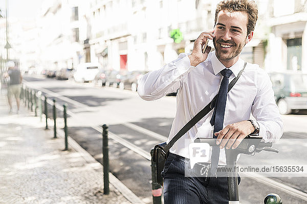 Lächelnder junger Geschäftsmann mit E-Scooter am Telefon in der Stadt  Lissabon  Portugal