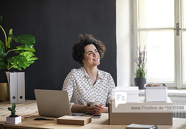 Lächelnde junge Frau sitzt am Schreibtisch mit Kaffeetasse,  Laptop und Paketen