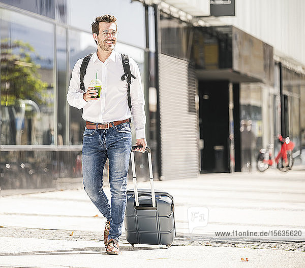 Lächelnder junger Mann mit Rollkoffer in der Stadt unterwegs