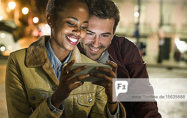 Portrait of happy young couple looking together at smartphone by night  Lisbon  Portugal