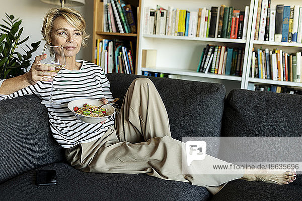 Reife Frau isst zu Hause hausgemachte Nudelgerichte und trinkt Wein auf der Couch