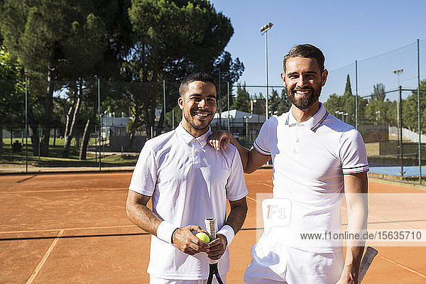 Two men in white sportswear holding rackets and smiling while standing on tennis court