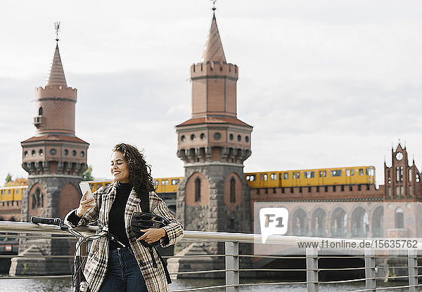 Lächelnde Frau mit Fahrrad mit Smartphone in der Stadt an der Oberbaumbrücke  Berlin  Deutschland