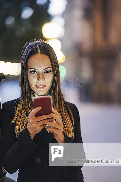 Portrait of smiling young woman leaning against lamp pole in the evening looking at cell phone