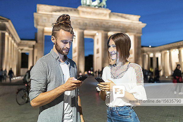 Happy couple using smartphones at Brandenburg gate at blue hour  Berlin  Germany